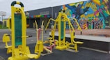 Outdoor gym equipment with vibrant yellow and pastel pink exercise machines set against a colorful mural wall featuring a bird and geometric patterns. In the background, there is a building with a multicolored sign and a basketball hoop on a court.