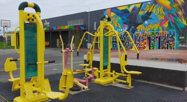 Outdoor gym equipment with vibrant yellow and pastel pink exercise machines set against a colorful mural wall featuring a bird and geometric patterns. In the background, there is a building with a multicolored sign and a basketball hoop on a court.