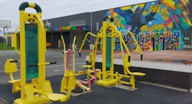 Outdoor gym equipment with vibrant yellow and pastel pink exercise machines set against a colorful mural wall featuring a bird and geometric patterns. In the background, there is a building with a multicolored sign and a basketball hoop on a court.