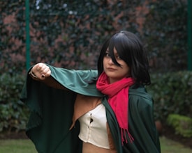 A person dressed in a cosplay outfit, featuring a green cape, black wig, and red scarf, stands in a garden setting. The individual is striking a dynamic pose with one arm extended forward.