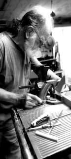 An elderly person with a white beard is concentrating on a task in a workshop setting. The individual is using a saw positioned on a workbench, surrounded by various tools. The environment appears to be cluttered with equipment and machinery.