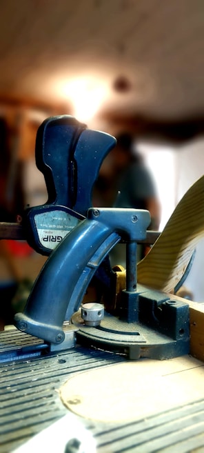 A close-up of a blue clamp tool positioned on a workbench, with a blurred figure and a bright light in the background. The tool is used for securing objects and appears to be in a workshop setting.