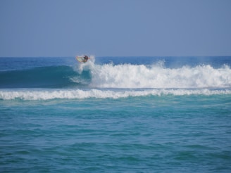A surfer skillfully rides a wave, catching air as they maneuver their board above the crest. The ocean is vibrant with shades of blue, and the waves create a sense of motion and energy.