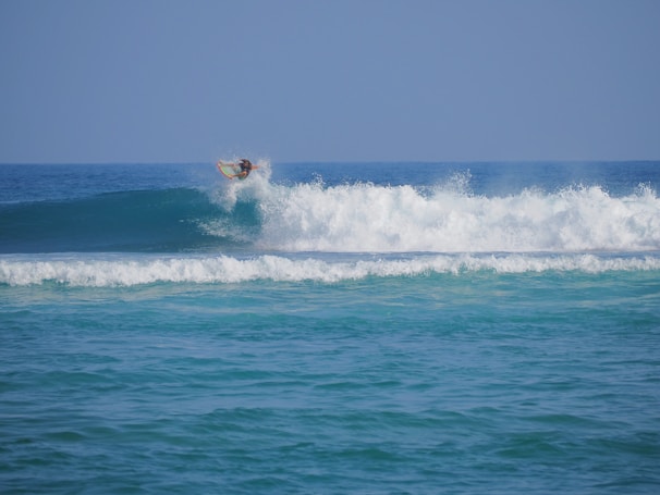 A surfer skillfully rides a wave, catching air as they maneuver their board above the crest. The ocean is vibrant with shades of blue, and the waves create a sense of motion and energy.