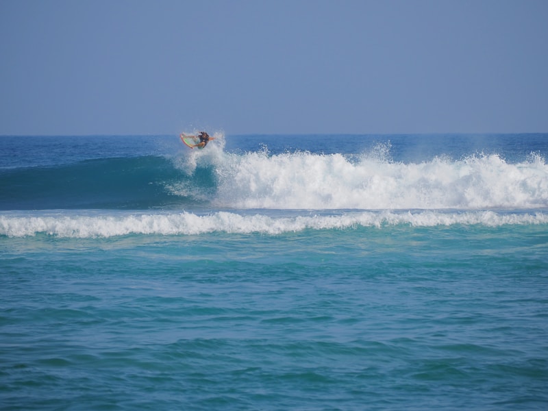 A surfer skillfully rides a wave, catching air as they maneuver their board above the crest. The ocean is vibrant with shades of blue, and the waves create a sense of motion and energy.