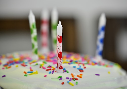Several colorful birthday candles are placed on a cake, which is topped with bright and varied sprinkles. The candles have designs of balloons and stripes, enhancing the festive atmosphere.