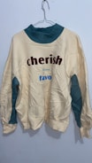 A wrinkled sweatshirt hanging on a wire hanger against a plain light-colored wall. The sweatshirt is primarily off-white with teal accents on the collar and underarm areas. It features the words 'cherish' in bold red letters and 'favo' in blue letters.