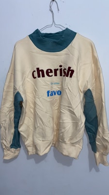 A wrinkled sweatshirt hanging on a wire hanger against a plain light-colored wall. The sweatshirt is primarily off-white with teal accents on the collar and underarm areas. It features the words 'cherish' in bold red letters and 'favo' in blue letters.