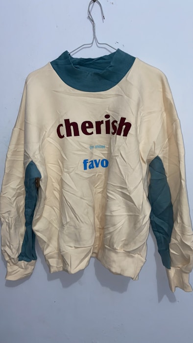 A wrinkled sweatshirt hanging on a wire hanger against a plain light-colored wall. The sweatshirt is primarily off-white with teal accents on the collar and underarm areas. It features the words 'cherish' in bold red letters and 'favo' in blue letters.