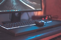 A close-up view of a computer setup featuring a large monitor with a visible desktop interface. The keyboard beneath it is illuminated with backlighting, and a wireless earbud case rests on the keyboard. The overall ambiance appears to be dimly lit, enhancing the glow from the screen and keyboard.