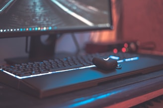 A close-up view of a computer setup featuring a large monitor with a visible desktop interface. The keyboard beneath it is illuminated with backlighting, and a wireless earbud case rests on the keyboard. The overall ambiance appears to be dimly lit, enhancing the glow from the screen and keyboard.