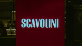 A red illuminated sign with the word 'SCAVOLINI' in bold white letters is positioned in front of what appears to be an interior setting, possibly a showroom or store. The background is dark, with some indistinct details, suggesting a dimly lit environment.