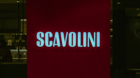 A red illuminated sign with the word 'SCAVOLINI' in bold white letters is positioned in front of what appears to be an interior setting, possibly a showroom or store. The background is dark, with some indistinct details, suggesting a dimly lit environment.