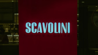 A red illuminated sign with the word 'SCAVOLINI' in bold white letters is positioned in front of what appears to be an interior setting, possibly a showroom or store. The background is dark, with some indistinct details, suggesting a dimly lit environment.