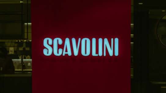 A red illuminated sign with the word 'SCAVOLINI' in bold white letters is positioned in front of what appears to be an interior setting, possibly a showroom or store. The background is dark, with some indistinct details, suggesting a dimly lit environment.