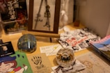 A tabletop display features various items including a framed artwork with '96 suke' written on it, a glass paperweight, business cards, a colorful illustrated page with a cartoon character, and black and white photographs with red text. The arrangement includes artistic elements and advertisements, suggesting a creative or promotional setting.