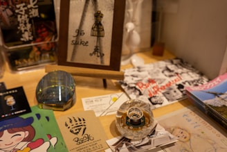 A tabletop display features various items including a framed artwork with '96 suke' written on it, a glass paperweight, business cards, a colorful illustrated page with a cartoon character, and black and white photographs with red text. The arrangement includes artistic elements and advertisements, suggesting a creative or promotional setting.