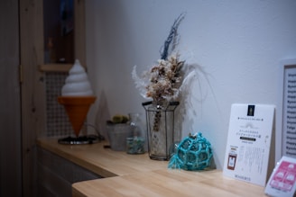 A cozy indoor setting featuring a wooden shelf adorned with decorative items, including a faux ice cream cone, a vase with dried flowers, a glass container with small objects, and a blue glass buoy. A menu or informational card is also displayed.