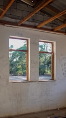 An interior view of a rustic, unfinished building with exposed wooden beams on the ceiling. There is graffiti on the white walls, and two large windows reveal a sunny, forested landscape outside.