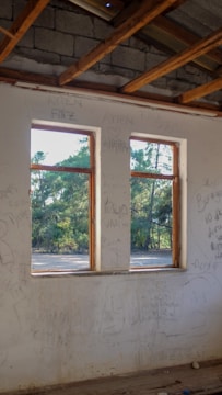 An interior view of a rustic, unfinished building with exposed wooden beams on the ceiling. There is graffiti on the white walls, and two large windows reveal a sunny, forested landscape outside.