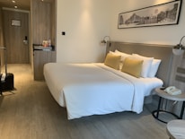 A neatly arranged hotel room featuring a large bed with white bedding and yellow pillows placed against a headboard. On one side of the room, there is a small nightstand with a telephone and a modern lamp. Opposite the bed, a wooden built-in closet and a door are visible. The room also includes a suitcase on the floor near the door. Above the bed, there is a black and white framed photograph on the wall.