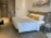 A neatly arranged hotel room featuring a large bed with white bedding and yellow pillows placed against a headboard. On one side of the room, there is a small nightstand with a telephone and a modern lamp. Opposite the bed, a wooden built-in closet and a door are visible. The room also includes a suitcase on the floor near the door. Above the bed, there is a black and white framed photograph on the wall.