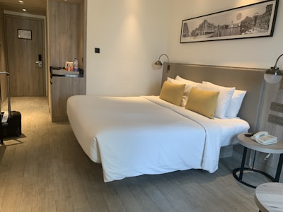 A neatly arranged hotel room featuring a large bed with white bedding and yellow pillows placed against a headboard. On one side of the room, there is a small nightstand with a telephone and a modern lamp. Opposite the bed, a wooden built-in closet and a door are visible. The room also includes a suitcase on the floor near the door. Above the bed, there is a black and white framed photograph on the wall.