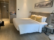 A neatly arranged hotel room featuring a large bed with white bedding and yellow pillows placed against a headboard. On one side of the room, there is a small nightstand with a telephone and a modern lamp. Opposite the bed, a wooden built-in closet and a door are visible. The room also includes a suitcase on the floor near the door. Above the bed, there is a black and white framed photograph on the wall.