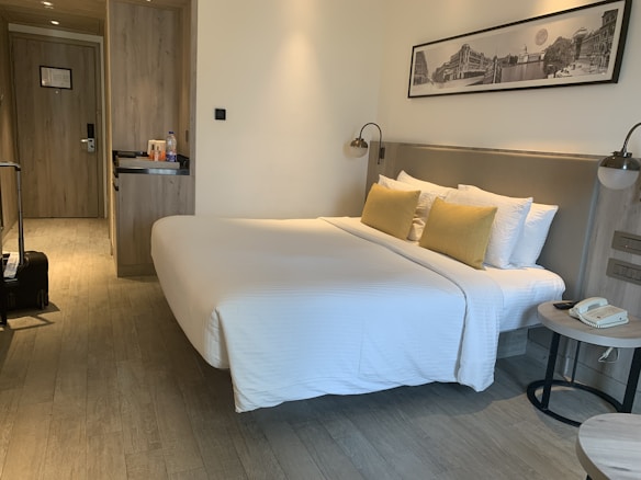 A neatly arranged hotel room featuring a large bed with white bedding and yellow pillows placed against a headboard. On one side of the room, there is a small nightstand with a telephone and a modern lamp. Opposite the bed, a wooden built-in closet and a door are visible. The room also includes a suitcase on the floor near the door. Above the bed, there is a black and white framed photograph on the wall.