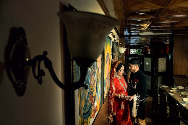 A couple is dressed in traditional attire, with the woman wearing a red saree and ornate jewelry, and the man in a black and gold sherwani. They stand close together in a warmly lit room with wooden furnishings and artistic paintings on the walls.