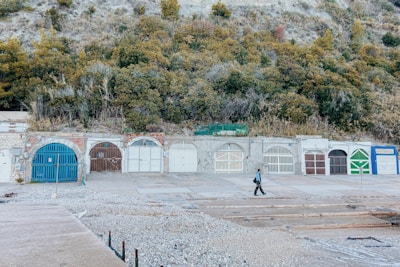 A row of closed, colored garage doors set against a stone wall with a grassy and rocky hill above. A person is walking across the concrete path in front of the garages.