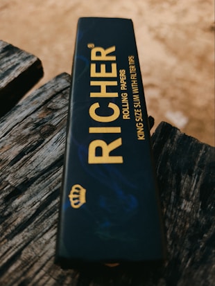 A package of rolling papers is placed on a textured, weathered wooden surface. The package is dark with gold lettering that reads 'RICHER' and includes details such as 'KING SIZE SLIM WITH FILTER TIPS'. There is also a small gold crown logo.