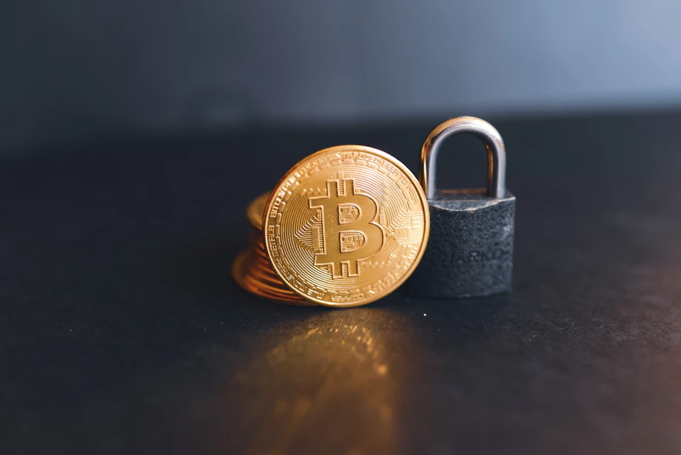 The image presents a scene set against a black background, with a gold-colored Bitcoin symbol prominently displayed on the left side. The Bitcoin symbol is surrounded by three coins, each varying in size and shape - one large coin positioned at the center, two smaller coins to its right, and another small coin located near the bottom of the image.

In the top right corner of the image, a silver lock can be seen, adding an element of mystery to the scene.