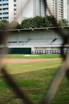 a baseball field through a fence with buildings in the background