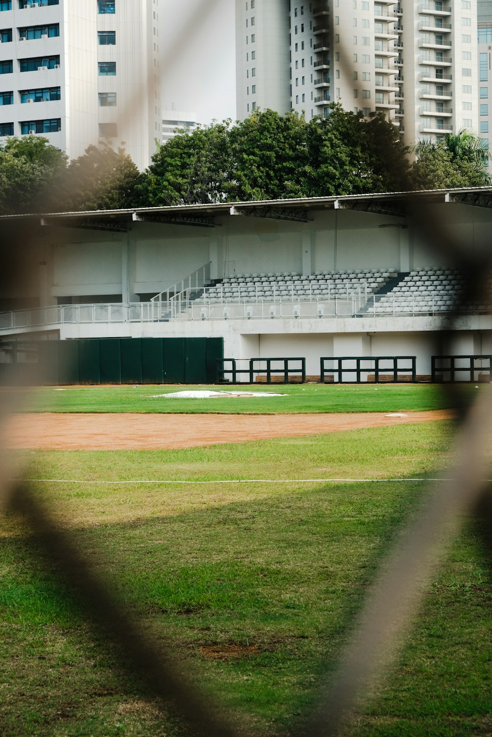 a baseball field through a fence with buildings in the background