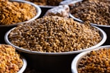 A variety of seeds or nuts displayed in large, round metal trays, arranged in a market setting. The seeds are pale brown and vary slightly in shade, suggesting different types or a mix.