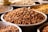 A close-up view of several metal trays filled with different types of unprocessed legumes, prominently featuring brown peanuts in the center. The trays are arranged closely together on a flat surface.