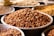 A close-up view of several metal trays filled with different types of unprocessed legumes, prominently featuring brown peanuts in the center. The trays are arranged closely together on a flat surface.