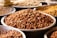 A close-up view of several metal trays filled with different types of unprocessed legumes, prominently featuring brown peanuts in the center. The trays are arranged closely together on a flat surface.