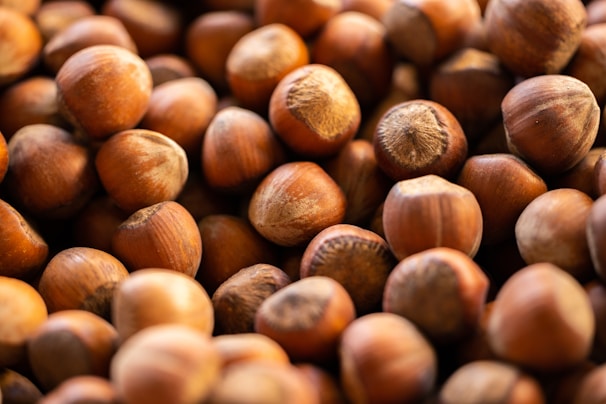 A close-up view of a large pile of hazelnuts with a focus on their rounded shapes and brown shells. The nuts are densely packed, displaying their textured surface and varied shades of brown.
