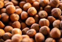 A close-up view of a large pile of hazelnuts with a focus on their rounded shapes and brown shells. The nuts are densely packed, displaying their textured surface and varied shades of brown.