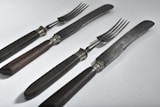 A set of vintage dining utensils featuring two knives and two forks with dark wooden handles. The blades and prongs are metal with a slightly tarnished appearance, suggesting age and use. The utensils are placed on a light, solid surface and arranged in a neat, parallel fashion.
