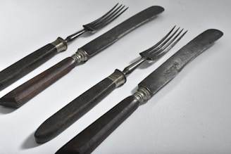 A set of vintage dining utensils featuring two knives and two forks with dark wooden handles. The blades and prongs are metal with a slightly tarnished appearance, suggesting age and use. The utensils are placed on a light, solid surface and arranged in a neat, parallel fashion.
