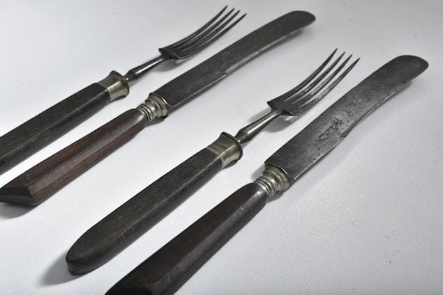 A set of vintage dining utensils featuring two knives and two forks with dark wooden handles. The blades and prongs are metal with a slightly tarnished appearance, suggesting age and use. The utensils are placed on a light, solid surface and arranged in a neat, parallel fashion.