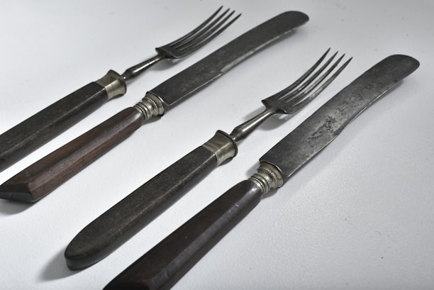 A set of vintage dining utensils featuring two knives and two forks with dark wooden handles. The blades and prongs are metal with a slightly tarnished appearance, suggesting age and use. The utensils are placed on a light, solid surface and arranged in a neat, parallel fashion.