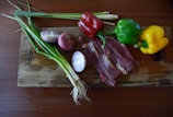 A wooden cutting board with various ingredients placed on it, including green onions, two types of onions, red, green, and yellow bell peppers, and slices of raw beef. The board is on a dark wooden table.