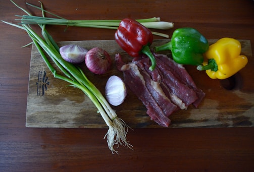 A wooden cutting board with various ingredients placed on it, including green onions, two types of onions, red, green, and yellow bell peppers, and slices of raw beef. The board is on a dark wooden table.