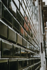 A close-up view of a tiled wall with glossy, dark green rectangular tiles arranged in a staggered pattern. The surface of the tiles reflects light, creating a shiny appearance. The perspective of the shot creates a sense of depth and leads the eye along the length of the wall.