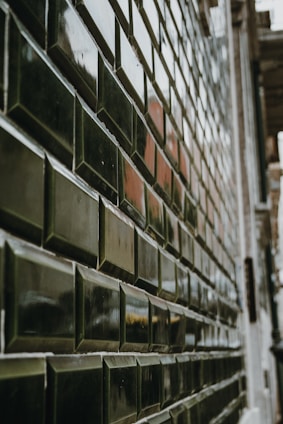 A close-up view of a tiled wall with glossy, dark green rectangular tiles arranged in a staggered pattern. The surface of the tiles reflects light, creating a shiny appearance. The perspective of the shot creates a sense of depth and leads the eye along the length of the wall.