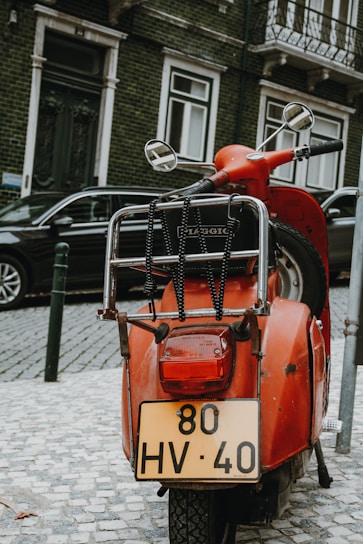 A sleek red Piaggio scooter parked on a cobblestone street with Italian architecture in the background.